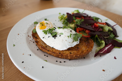 Poached egg on a piece of bread with green salad on a plate and on the wooden table. Healthy Breakfast