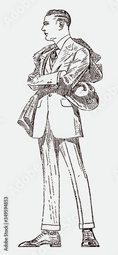 Standing young man from the early 20th century in a suit, with arms crossed, holding a hat in his hand and wearing a coat over his shoulder