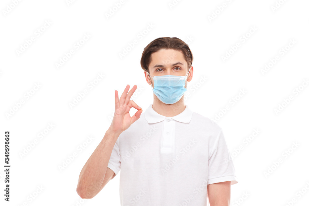 A man in a white T-shirt of European appearance in a medical mask, shows a thumb up. In the mask you are protected from covid 19 viruses.