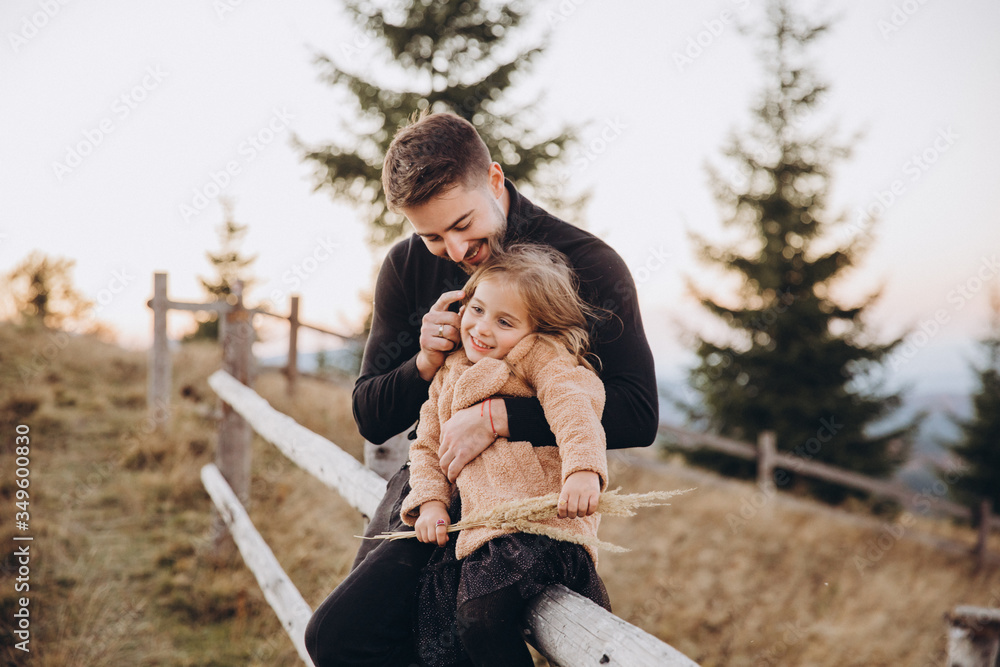 Stylish young family in the autumn mountains. Dad and daughter are sitting on the fence and hugging against the background of the forest and mountain peaks at sunset. Stylish young family in the autumn mountains. Dad and daughter are sitting on the fence and hugging against the background of the forest and mountain peaks at sunset.