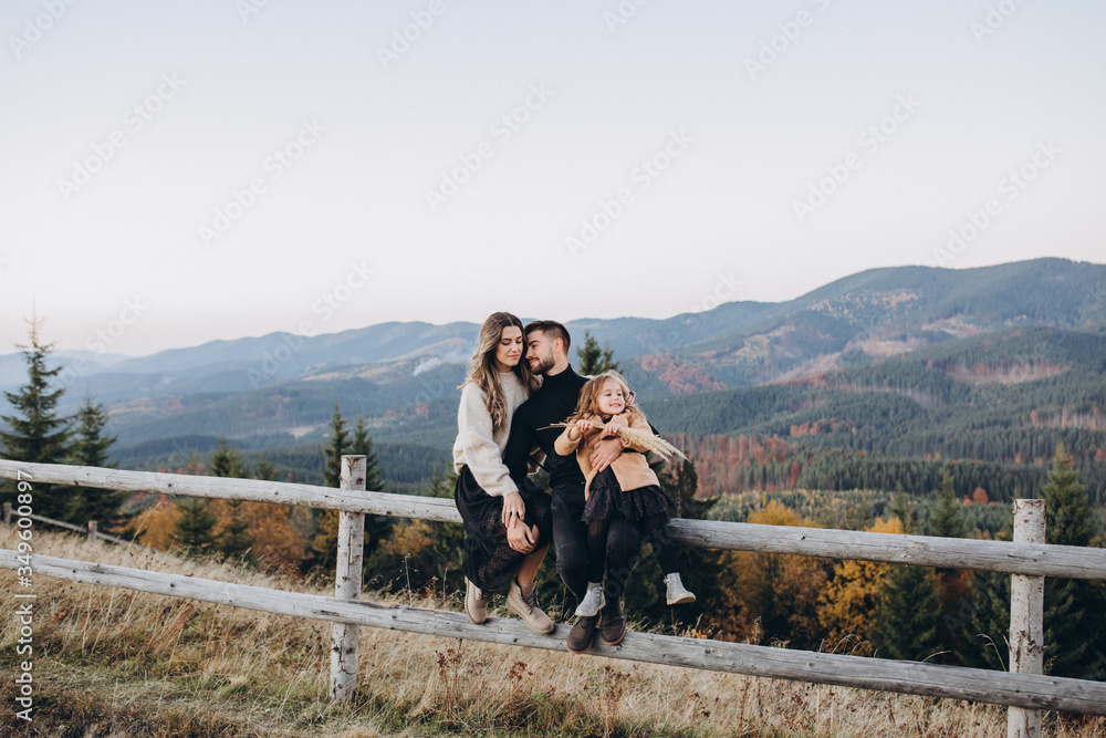 Stylish young family in the autumn mountains. A guy and a girl, together with their daughter, are sitting on a fence amid a forest and mountain peaks at sunset.