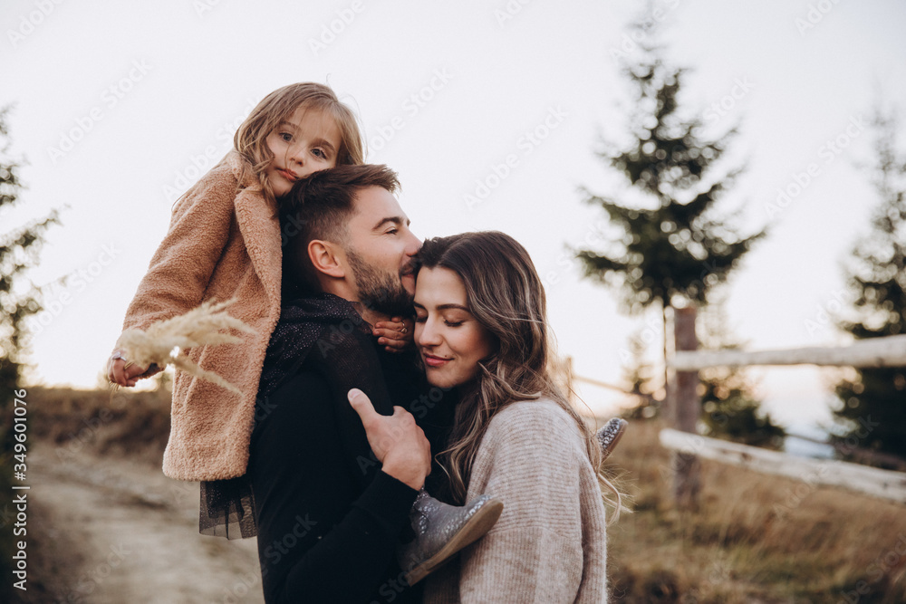 Fototapeta premium Stylish young family in the autumn mountains. A guy and a girl together with their daughter, are standing and hugging on the road against the background of a fence, forest and mountain peaks at sunset