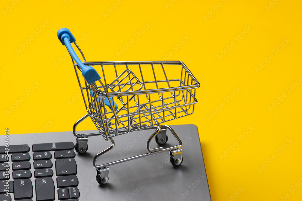 Global online supermarket. Shopping cart on laptop keyboard. Yellow studio background