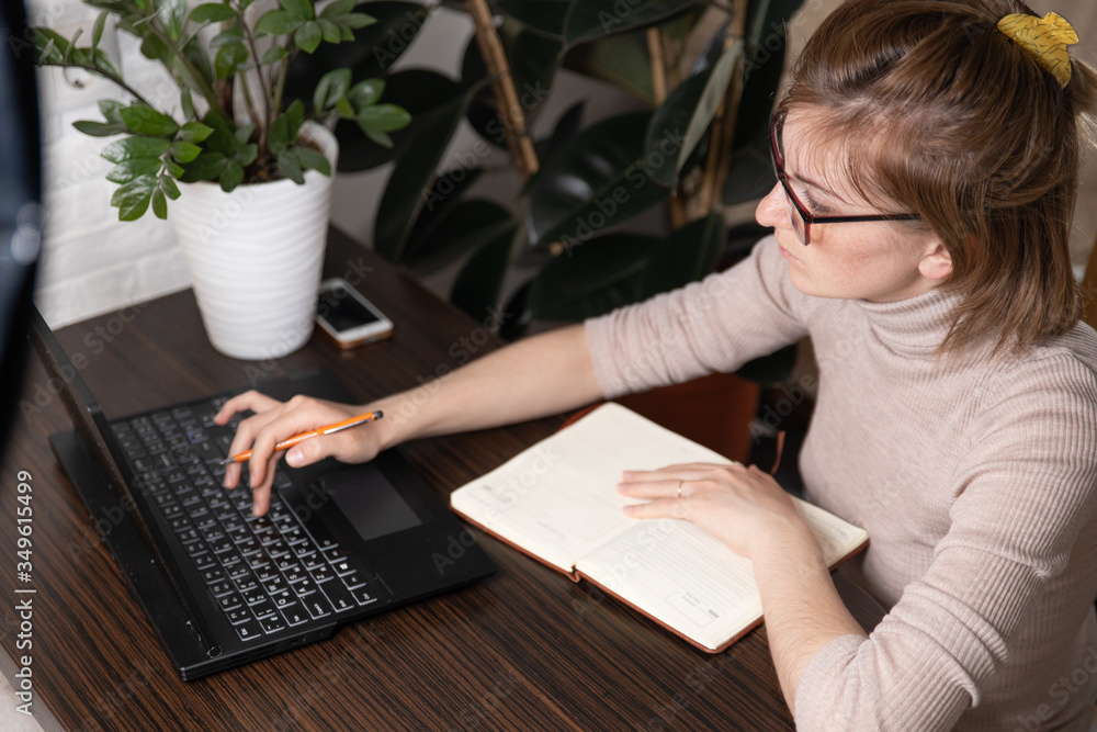 Side view. Young business woman sitting at table and taking notes in notebook.On table is laptop, smartphone. On computer screen graphics and charts. Student learning online. Blogger. Side view. Young business woman sitting at table and taking notes in notebook.On table is laptop, smartphone. On computer screen graphics and charts. Student learning online. Blogger.