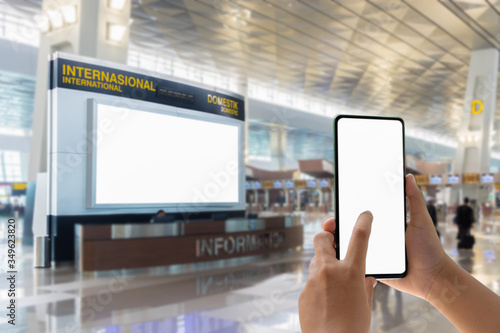 Woman passenger hand holding empty screen of smart phone and check-in counter airport terminal background.