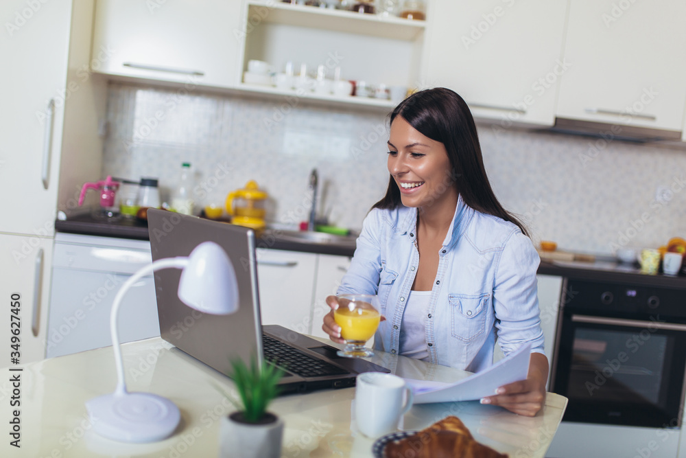 Attractive young woman working with laptop computer and documents while sitting at the kitchen.