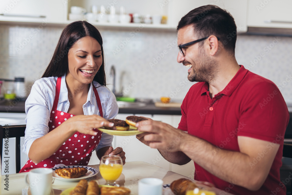 Cheerful young couple having breakfast in kitchen eating donuts.