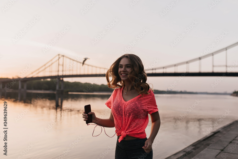 Spectacular girl with smartphone running near river in evening. Outdoor shot of laughing pretty female model enjoying training.