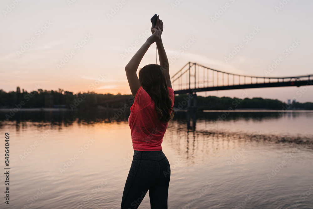 Fototapeta premium Enchanting shapely girl standing with hands up and looking at river. Outdoor photo from back of amazing female model relaxing after training near lake.