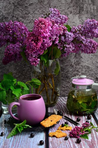 Vertical composition of a cup of herbal tea, a teapot, cookies and a bouquet of lilacs in a glass vase on a dark background. Spring still life.