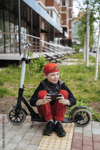 A teenage boy sits next to a scooter, playing on the phone