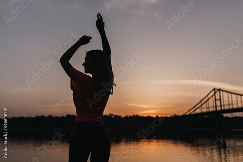 Portrait from back of graceful girl standing on nature background in evening. Outdoor shot of inspired lady relaxing after training and looking at sunset.