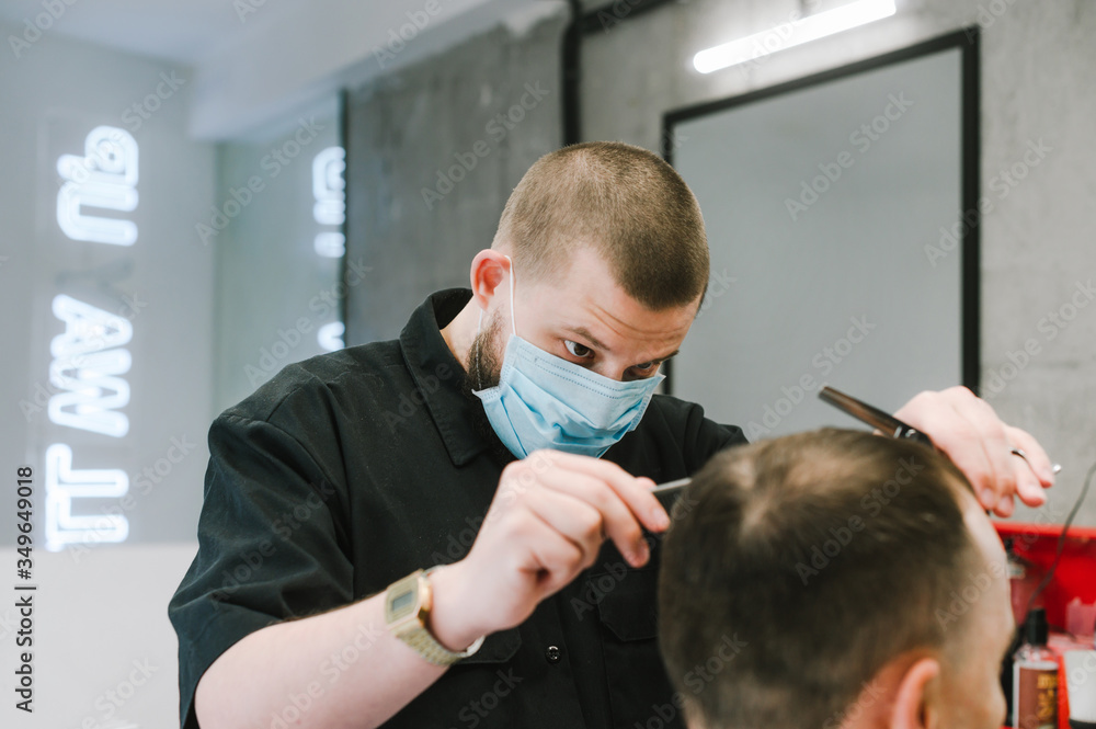 Φωτογραφία Professional barber in a medical mask cuts the client with scissors and a comb in a men's hairdresser with a concentrated face
