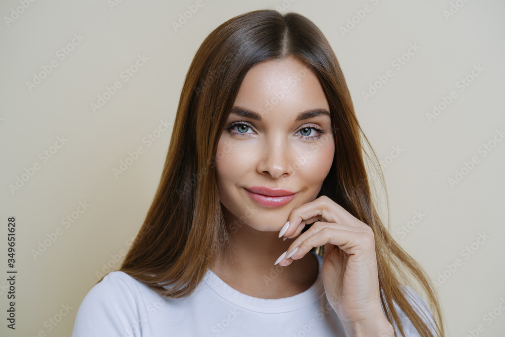 Charming good looking millennial woman keeps hand near face, has healthy glowing skin, dark hair, blue eyes, dressed in casual wear, isolated over brown background. Human face expressions concept Charming good looking millennial woman keeps hand near face, has healthy glowing skin, dark hair, blue eyes, dressed in casual wear, isolated over brown background. Human face expressions concept
