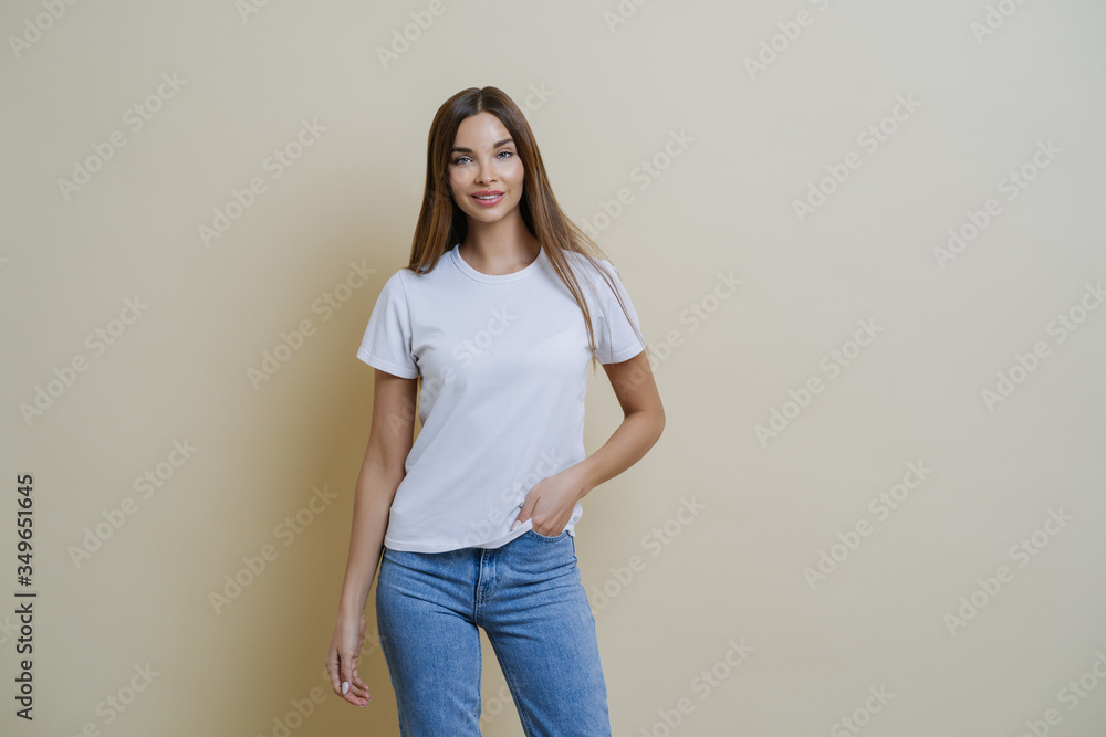 Studio shot of pretty slim woman keeps hand in pocket, wears casual t shirt and jeans, stands in relaxed pose, looks at camera self confident, isolated over beige background talks casually with client Studio shot of pretty slim woman keeps hand in pocket, wears casual t shirt and jeans, stands in relaxed pose, looks at camera self confident, isolated over beige background talks casually with client