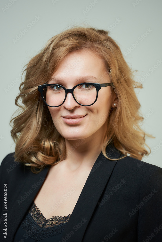 A stylish young woman in glasses dressed in a black suit with a black shirt. A close-up portrait of a blond lady with a smile who wears a business outfit. A concept of an office style wardrobe.