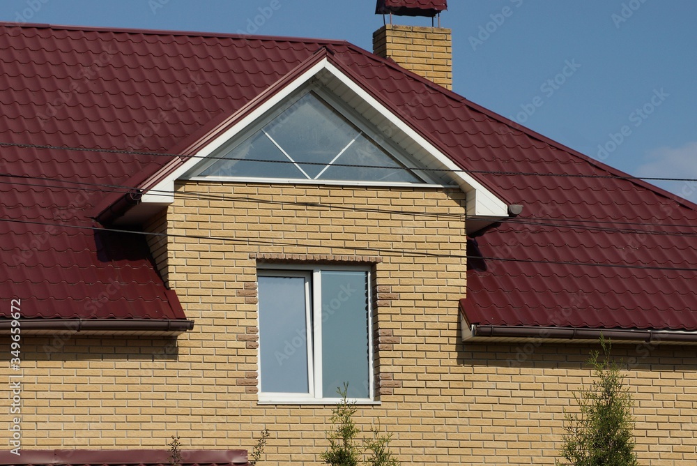 facade of a brown brick private house with a white window under a tiled roof against a blue sky