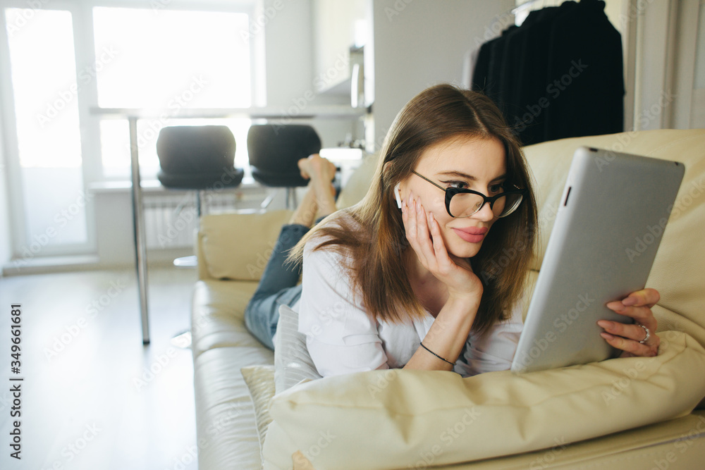 Woman in сasual jeans and white shirt on sofa at home browsing internet from digital tablet, so comfortable, so nice light design interior indoors Woman in сasual jeans and white shirt on sofa at home browsing internet from digital tablet, so comfortable, so nice light design interior indoors