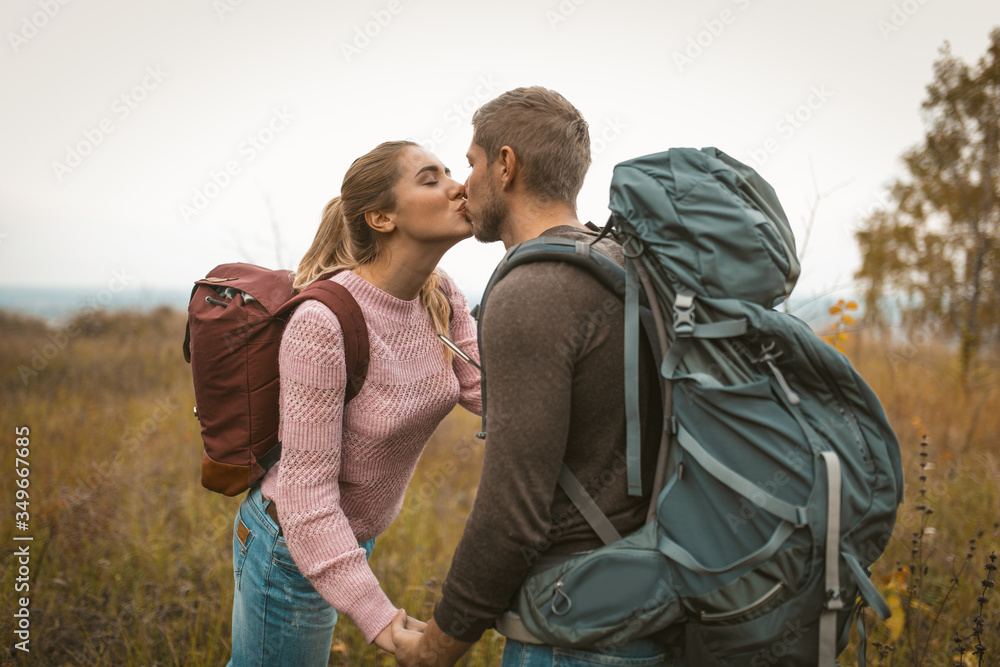 Loving backpackers kiss in the field outdoors. Happy couple of travelers, young man and woman standing against the backdrop of autumn nature. Love for adventure concept