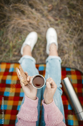 Picnic or snack in nature. Female hands holding a cup of tea and a sandwich. Young woman or teen girl snacking while sitting on grass. Close-up shot. High angle view