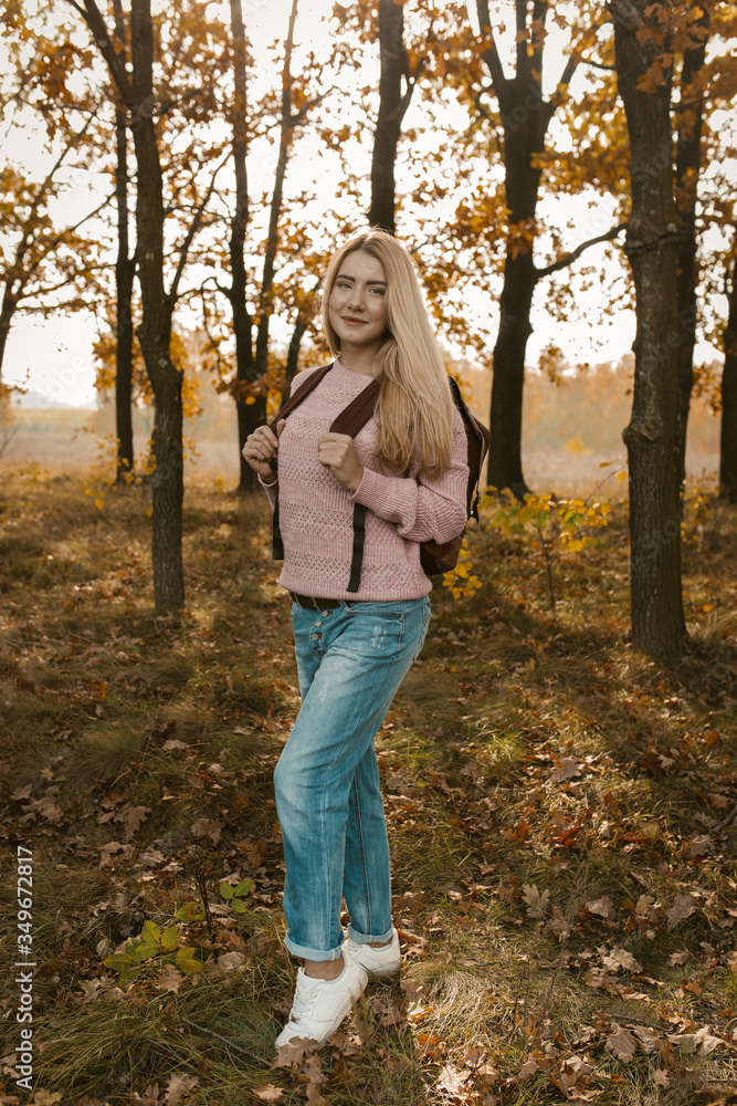 Female traveler posing with a backpack on the background of the autumn forest backlit by the sun's rays. Beautiful blonde travels in nature. Hiking concept Female traveler posing with a backpack on the background of the autumn forest backlit by the sun's rays. Beautiful blonde travels in nature. Hiking concept