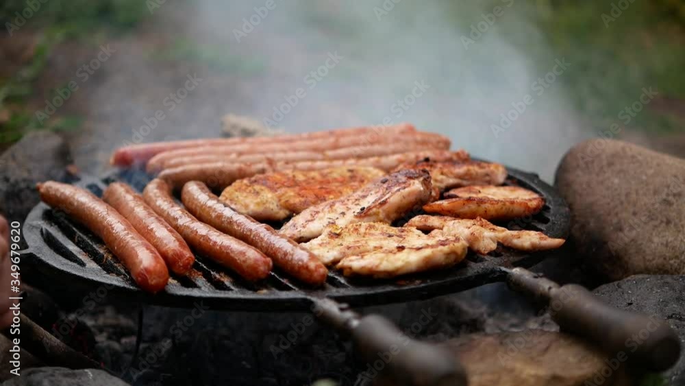 Sausages and chicken meat on the grill, grillingchicken and sausages on the grill in the yard, hands raise the grill and set the charcoal grill Sausages and chicken meat on the grill, grillingchicken and sausages on the grill in the yard, hands raise the grill and set the charcoal grill