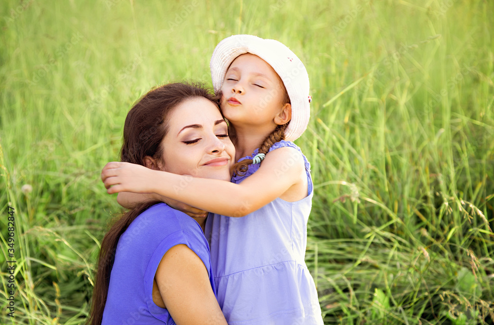 Beautiful mother hugging and kissing her cute small daughter on summer green grass background. Closeup Beautiful mother hugging and kissing her cute small daughter on summer green grass background. Closeup