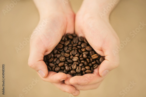 A person is holding a large handful of quality aromatic roasted coffee beans on a beige background, illuminated by light.