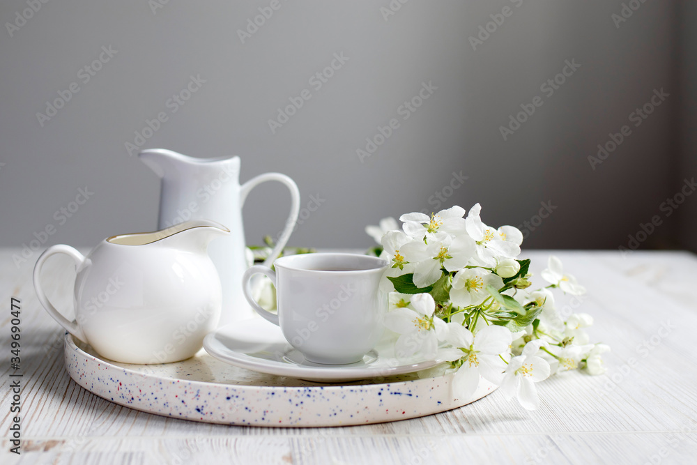 Vase with a blossoming branch of an apple tree. A set of white dishes: a cup of coffee, milk jug, a jug, a tray on the table against the background of a blue wall. Vase with a blossoming branch of an apple tree. A set of white dishes: a cup of coffee, milk jug, a jug, a tray on the table against the background of a blue wall.