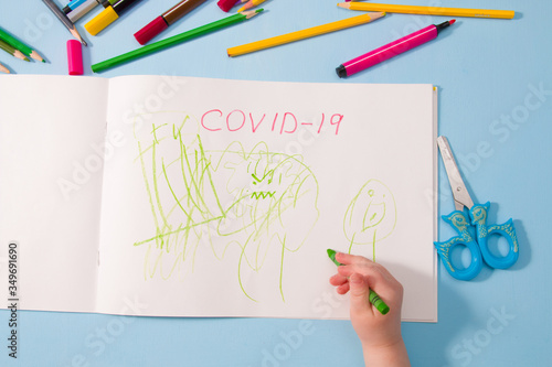 a small child draws green wax crayon in an album coronavirus, pencils on a blue background, top view, copy space, activities with children at home during quarantine
