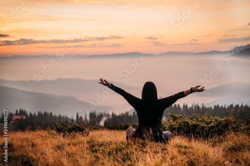  Yoga woman sitting on top of a mountain at sunrise. Mountains in the fog. The view from the back. arms around. Meditation. The balance of psychological life. Appeasement. Peace and joy in the soul