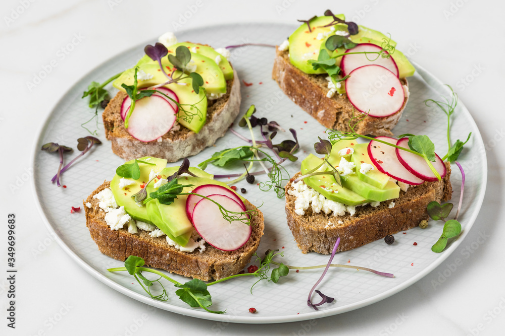 Healthy avocado toasts with radish, feta cheese, pea sprouts and pepper in a plate on white marble table