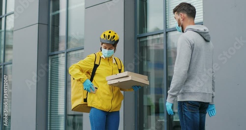 Caucasian young female courier in casque, gloves and medical mask calling to client on phone and giving pizza to man at street. Woman byciclist with bag bringing boxes to male customer outdoors. photo