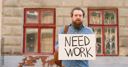 Portrait shot of unemployed Caucasian young man standing on street in town and showing poster with Need Work words. Male workless demonstrating board with announcement. Jobless person after lockdown. photo