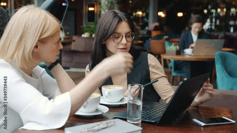 Female business partners attractive young and mature women are talking pointing at laptop screen during negotiations in restaurant. Businesswoman and work concept. Female business partners attractive young and mature women are talking pointing at laptop screen during negotiations in restaurant. Businesswoman and work concept.
