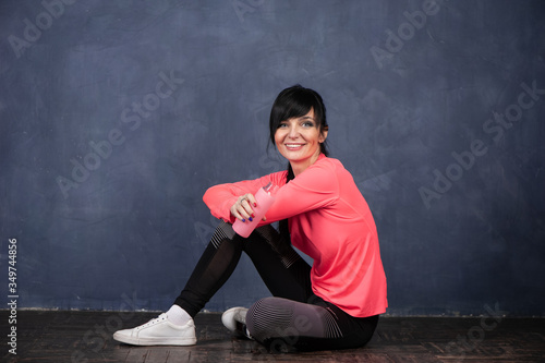 Trendy beautiful girl, brunette in bright sports clothes sits on the floor and looks into the camera, holding a bottle of water. Ready to start training. The concept of sports, fitness, space for text