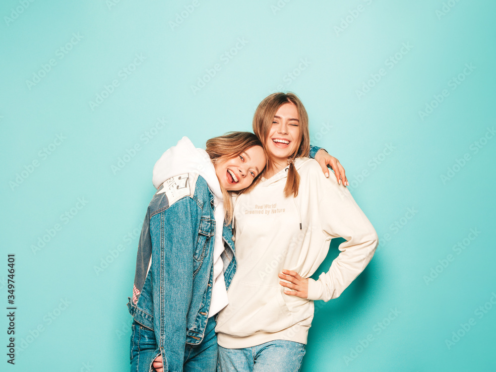 Two young beautiful brunette smiling hipster girls in trendy summer hoodie and jeans jacket clothes. Sexy carefree women posing near blue wall. Trendy and positive models having fun Two young beautiful brunette smiling hipster girls in trendy summer hoodie and jeans jacket clothes. Sexy carefree women posing near blue wall. Trendy and positive models having fun