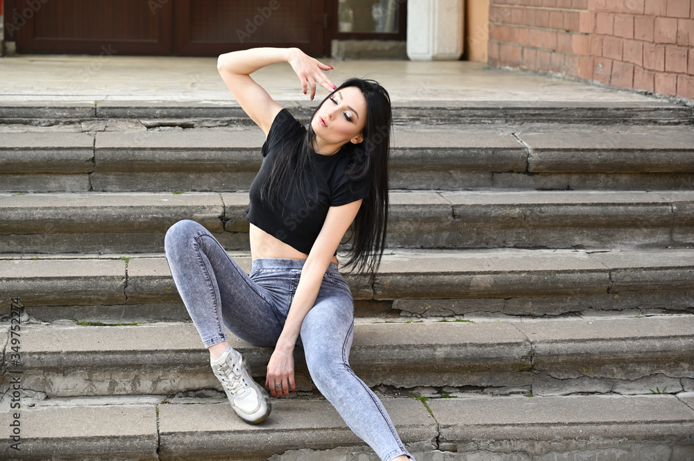 Fashionable model sits on the stairs of the building. Full-length portrait in spring weather outdoors, caucasian brunette girl with a slim figure in jeans and a black T-shirt. Fashionable model sits on the stairs of the building. Full-length portrait in spring weather outdoors, caucasian brunette girl with a slim figure in jeans and a black T-shirt.