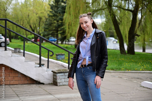 Portrait of a young pretty girl with a smile in a black jacket and jeans. Model posing standing in spring park outdoors in the city.