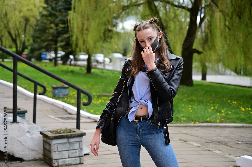 Model posing standing in spring park outdoors in the city. Photo of a young pretty girl in a protective mask from the virus in a black jacket and jeans.