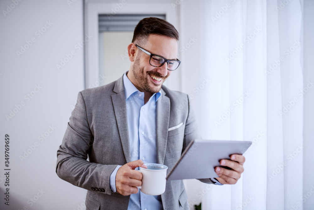 Attractive handsome classy caucasian smiling businessman in suit and with eyeglasses using tablet and holding mug with coffee while standing next to window in his office. Attractive handsome classy caucasian smiling businessman in suit and with eyeglasses using tablet and holding mug with coffee while standing next to window in his office.