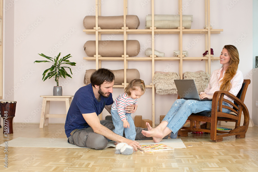 Mother working from home on her laptop while her daughter playing on the floor with father Mother working from home on her laptop while her daughter playing on the floor with father
