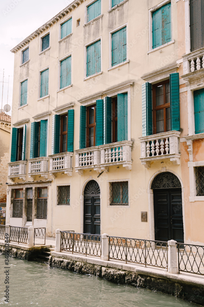 Closeup of the facade of a building, on the streets of Venice, Italy. The four-story building is sand-colored with green forged wooden shutters and balconies with white columns