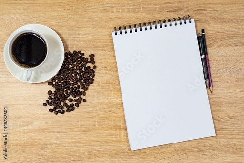 on the table there is a mug with aromatic coffee, coffee grains near a cup on the surface, a pencil and pen next to a notebook, a sheet of paper for writing ideas © Aleksandr Lavrinenko