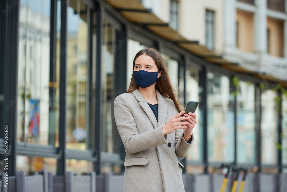 Fototapeta premium A girl with long hair in a navy blue face mask to avoid the spread coronavirus uses a smartphone in the street. A woman in a face mask against COVID-19 wears a coat looks away in the city.