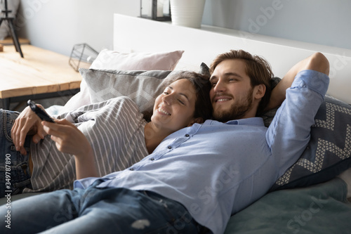 Happy young Caucasian man and woman lying relaxing on comfortable bed at home watching TV together, smiling millennial couple rest in bedroom, enjoy lazy leisure weekend indoors, relaxation concept