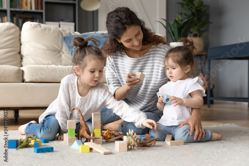 Loving young Caucasian mother sit on floor in living room playing building bricks with little daughters  caring mom or nanny construct with wooden block with small girls children at home together