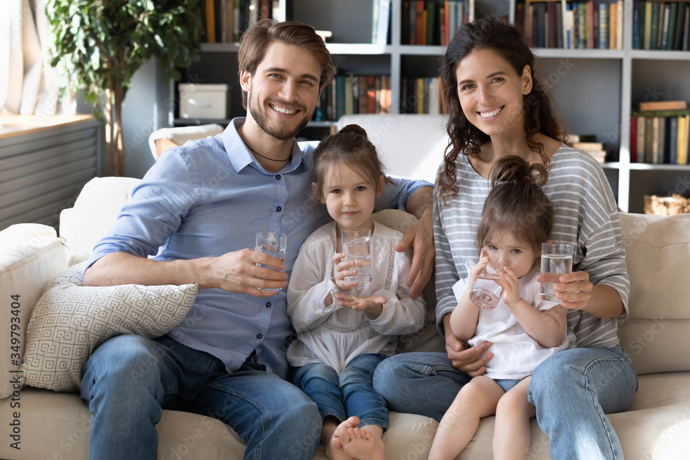 Portrait of happy young Caucasian family with little daughters on couch at home recommend drinking water, smiling parents with small girls children follow healthy lifestyle, dieting, wellness concept Portrait of happy young Caucasian family with little daughters on couch at home recommend drinking water, smiling parents with small girls children follow healthy lifestyle, dieting, wellness concept