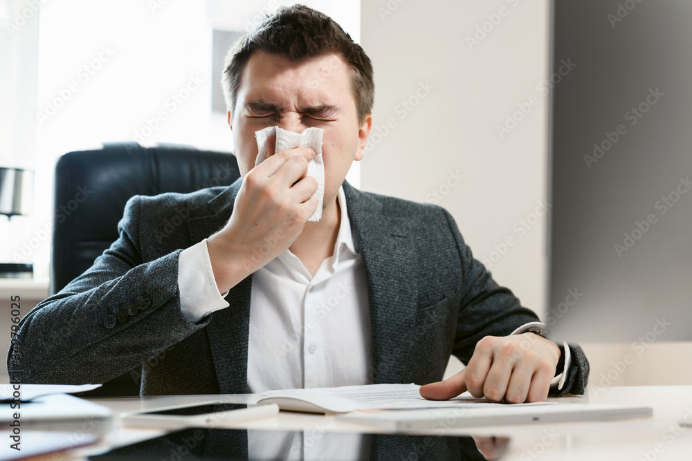 A young businessman or office worker sneezes, coughs, blows his nose at the workplace. Overworked worker got colds and flu. High temperature with ARVI or SARS. Epidemic of influenza or coronavirus. A young businessman or office worker sneezes, coughs, blows his nose at the workplace. Overworked worker got colds and flu. High temperature with ARVI or SARS. Epidemic of influenza or coronavirus.