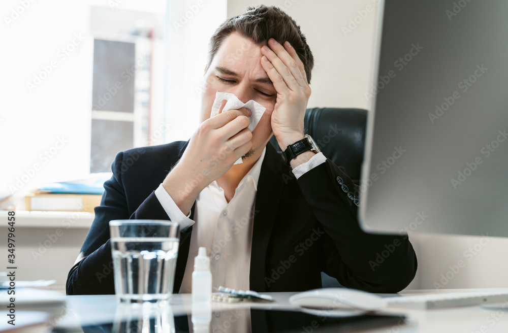 A young businessman or office worker sneezes, coughs, blows his nose at the workplace. Overworked worker got colds and flu. High temperature with ARVI or SARS. Epidemic of influenza or coronavirus. A young businessman or office worker sneezes, coughs, blows his nose at the workplace. Overworked worker got colds and flu. High temperature with ARVI or SARS. Epidemic of influenza or coronavirus.